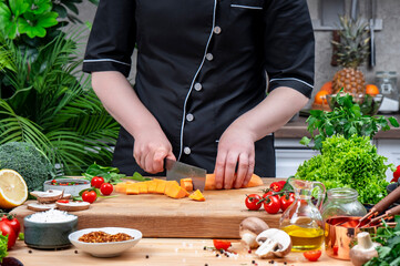 Chef cuts organic sweet potato into cubes with nakiri knife.. Cozy kitchen with wooden table, kitchenware, vegetables, herbs and ingredients for cooking. Healthy vegan eating, culinary, recipes