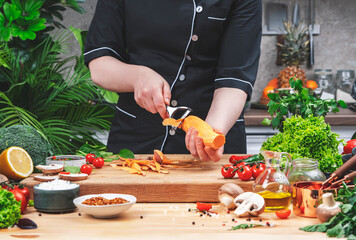 Home cook peels sweet potato with vegetable peeler. Cozy kitchen with wooden table, kitchenware, vegetables, herbs and ingredients for cooking. Healthy vegan eating, culinary, recipes, food blogging 