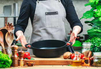 Chef holds empty cast iron saucepan in hands. Cozy kitchen with wooden table, kitchenware, vegetables, herbs and ingredients for cooking. Healthy eating, culinary, recipes, food blogging