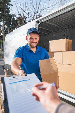 Smiling delivery man handing package and clipboard for signature near a van with stacked boxes. Concept of delivery person and package delivery.