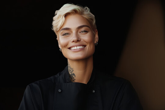 Chef with short blonde hair smiles confidently while wearing a black chef coat and showcasing tattoos