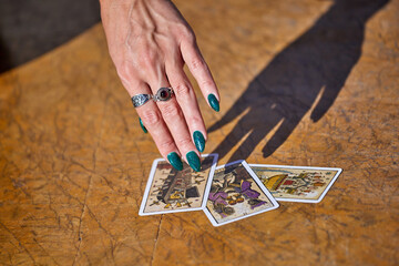 A woman's hand with green glittery nails touches three tarot cards on a wooden table; she wears two...