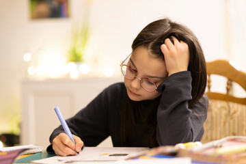 Little dark-haired girl in glasses sitting in the room at a wooden table reading a book. Blurred background. Children's educational concept.