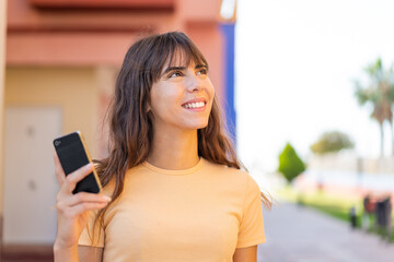 Fototapeta premium Young woman using mobile phone at outdoors looking up while smiling