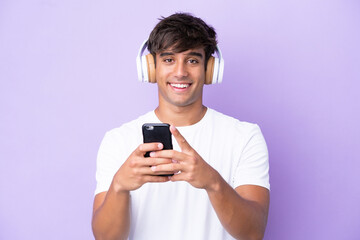 Fototapeta premium Young caucasian man isolated on purple background listening music with a mobile and looking front