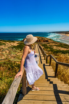 Mature woman tourist admiring picturesque view to cliff, ocean and sandy beach standing on wooden promenade in summertime. Side view. Bordeira beach panorama in Portugal