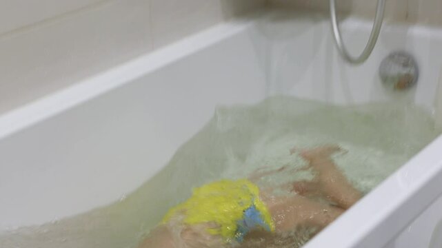 young boy in swimming goggles and a yellow bathing suit submerges himself underwater in a bathtub, learning to hold his breath while taking a bath.