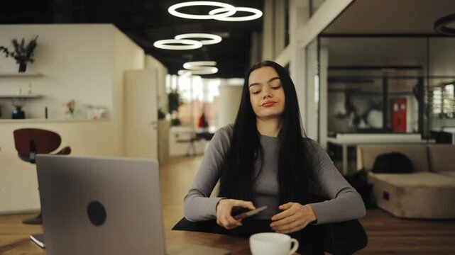 A beautiful woman is bored in a beautiful office while working on a laptop, picks up the phone to distract herself, relax, break at work, remote work, lack of concentration, cellphone, typing