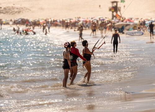 Female kitesurfing instructor teaching kite balancing techniques to a mother and her son on a crowded beach in Tarifa, Spain, on a sunny day.
