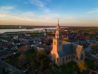Fototapeta premium Aerial view of a Dutch Catholic church on the banks of the De Lek river in the southern Netherlands. Spring landscape with houses clustered on the vast plain of the Netherlands in the evening at sunse