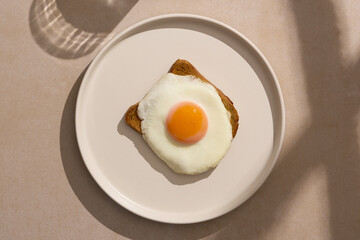 Sunny side up egg with vibrant yolk perched on toasted bread aon white plate, ready to eat. Minimal design, top view, direct morning sunlight with beautiful window and glass shadows. 