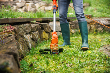 Grass trimmer. Woman gardener is cutting grass next stone wall in garden. Lawn care