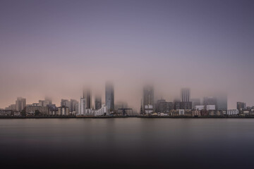 Obraz premium Canary Wharf shot during a misty day at sunset, allowing the colours of the sky diffuse and shroud the cityscape in the golden hour.