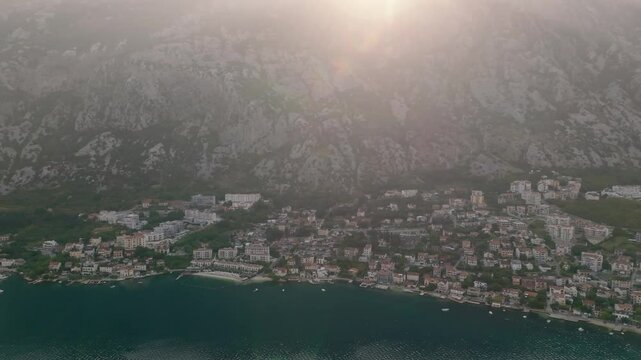 Cinematic drone view of the coastal town of Dobrota, Kotor, with residential houses at sunrise, mountains, and the sun over the peaks, Montenegro, Europe