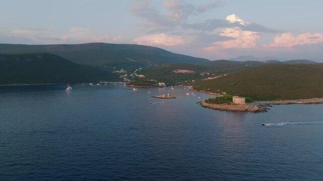 Cinematic drone flyover to the island of Vavednja, Zanjica or Mala Gospa with the Manastir Zanjica fortress over a passing boat, featuring the Fort Arza, Lustica Bay, and mountains in the background