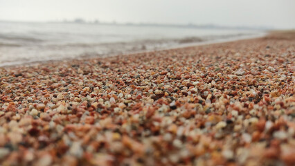 A detailed close-up of multicolored pebbles covering a beach, with the ocean waves softly blurred in the background. The mixture of warm and cool tones creates a natural, textured landscape.