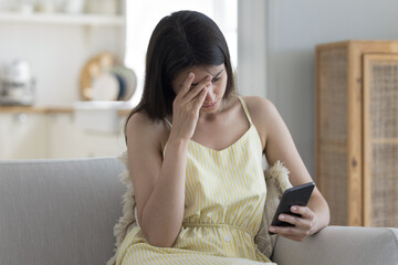Bad news. Distressed confused teen girl sit on couch with hand on head hold cellphone overwhelmed...