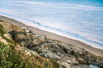 A beach with a rocky cliff and a body of water. The water is blue and the sky is pink