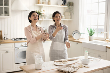 Cooking can be so funny. Joyful family two diverse age females senior mom adult kid in aprons bake sweet buns in modern home kitchen laughing having fun juggling pieces of dough in playful crazy mood