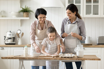 Funny family cooking. Happy three female generations mature grandmother young mother little kid daughter in aprons gather at home kitchen table to cook dessert bake pie make dough for homemade bakery