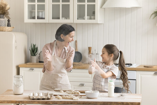 Crazy culinary. Joyful laughing senior grandmother playing having fun in kitchen with excited little grandkid girl scream in delight making snow from flour when kneading rolling dough for baking pies