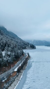Ice-covered lake at the foot of the steep mountains overgrown with evergreen pine-tree woods. A highway is on the lakefront close to water. Aerial view. Vertical video.