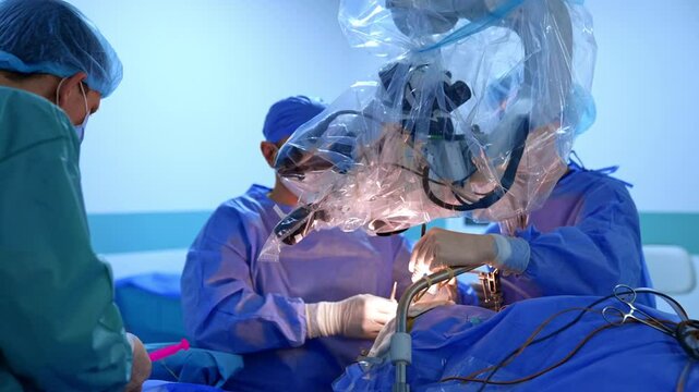 Two neurosurgeons sit behind the microscope above the operating table. Male assistant arranges tools at the surgical stand.