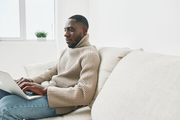 A young Black man in a cozy beige sweater working on a laptop while sitting on a light sofa, showcasing focus and productivity at home