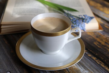 A white cup with a golden rim stands on the table with an open book and flowers on the book.
