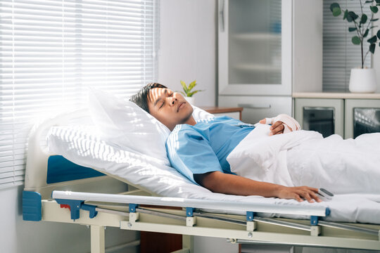Bandaged asian male patient lying in hospital room, healing after surgical procedure, soft sunlight filtering through window blinds