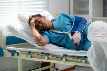 Young Asian male patient lying in hospital bed, wearing hospital gown, resting with broken arm in...