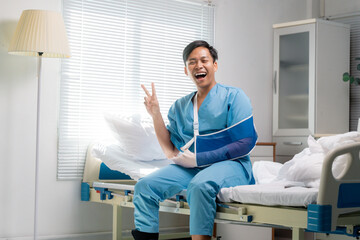Smiling patient in medical gown resting on hospital bed, wearing arm cast and gesturing victory...