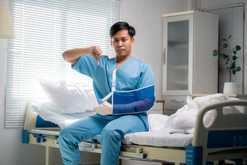 Young asian medical professional in blue scrubs and arm sling showing thumbs down, signaling...