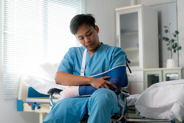 Asian doctor wearing blue scrubs sitting on wheelchair with arm sling and bandaged hand recovering...
