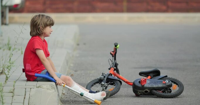 Boy with broken leg in gypsum plaster cast and medical orthopedic crutches sits on sidewalk border curb near bike, feeling sorry for being not able to take a ride