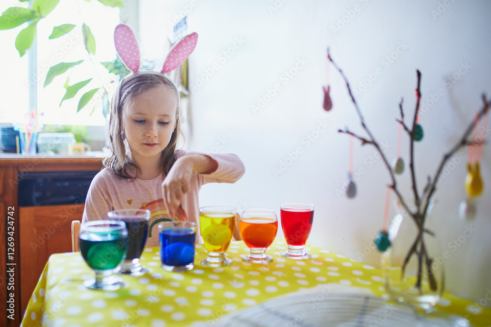 Wall mural Adorable preschooler girl dyeing Easter eggs at home. Child painting colorful eggs for Easter hunt. Kid getting ready for Easter celebration. Family traditions