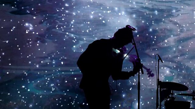 Music. Silhouette of a violinist playing a glowing electronic violin on the background of a large screen. Hand held.