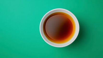 Overhead shot showcases a white bowl filled with dark brown liquid, possibly sauce or broth, set against a flat, solid green background. The scene is