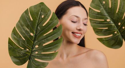 Beautiful smiling woman posing with tropical monstera leaves on a beige. Beauty concept.