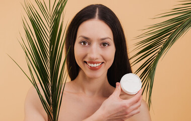 Beautiful smiling woman holding cosmetic cream with tropical palm leaves on beige background