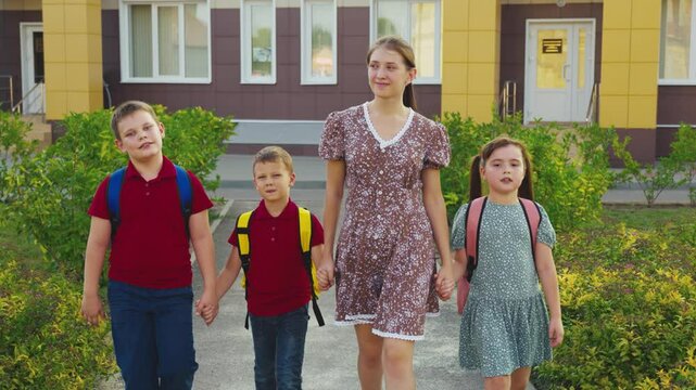 Mother leading children from school, female mother holding children hand, happy family, young nanny with children, schoolchildren with backpacks walking with aunt by the hand, little boys girl walking