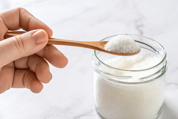 Woman fingers hold wooden spoon full of monosodium glutamate white crystals over glass jar. Favor enhancer MSG, food additive E621 concept.