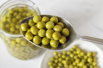 Full spoon of canned green peas close-up. Marinated sweet peas in a metal spoon over jar and plate. Snack, salad ingredient, vegetable protein, vegetarian food.