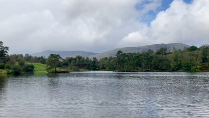 Tarn Hows in the Lake District National Park is seen on a partly cloudy day with a tranquil lake, forest, and mountains - Cumbria, UK