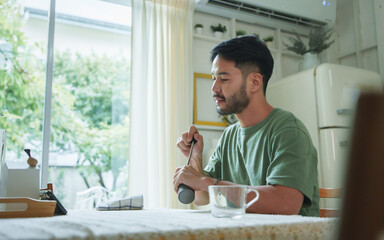 Asian man in green T-shirt sitting at a dining table grinding coffee beans while watching a smartphone in a dining room with natural light from large windows.