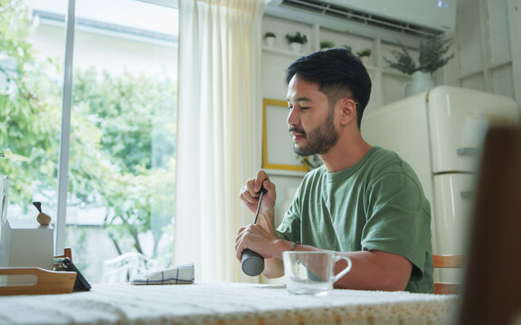 Asian man in green T-shirt sitting at a dining table grinding coffee beans while watching a smartphone in a dining room with natural light from large windows.