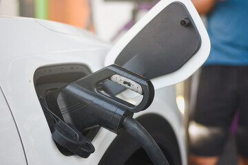 the side of a white electric vehicle with the charging port open and a charging cable plugged into it. The charging port is located near the front of the vehicle, just behind the side mirror.