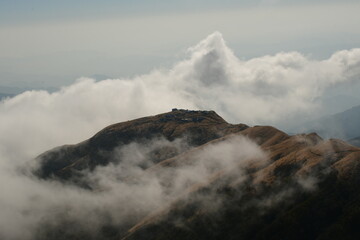 A mesmerizing view of a remote mountain settlement sitting atop a golden-hued ridge, partially enveloped by misty clouds. The contrast between the rugged landscape