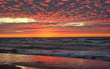 Fototapeta premium An intense sunset with deep orange and red hues reflecting over Lake Michigan. The waves crash along the shore as the sky’s cloud patterns add depth and texture to the scene.