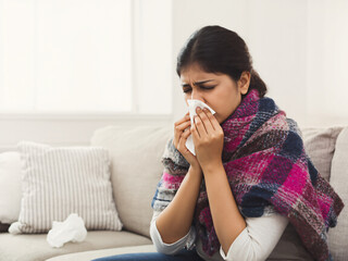 A woman wrapped in a warm blanket, sneezing into a tissue while suffering from a cold or flu, symbolizing seasonal illness, home recovery, and self-care.